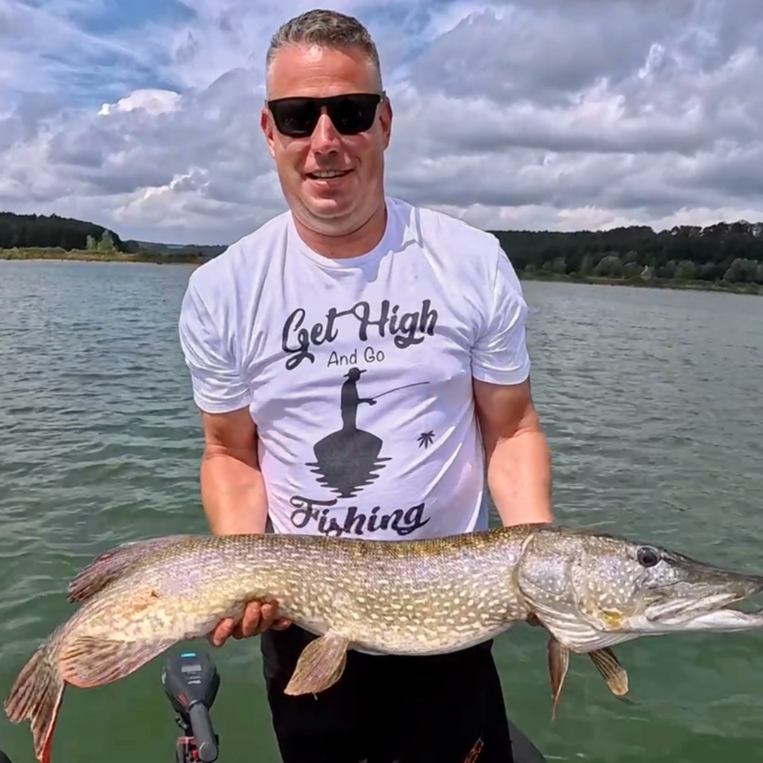 Angler auf dem Boot mit großem Hecht – trägt „Get HIGH and go FISHING“ T-Shirt, sonniger Tag auf dem See, Outdoor Fishing Motiv für Wels- und Raubfischangler