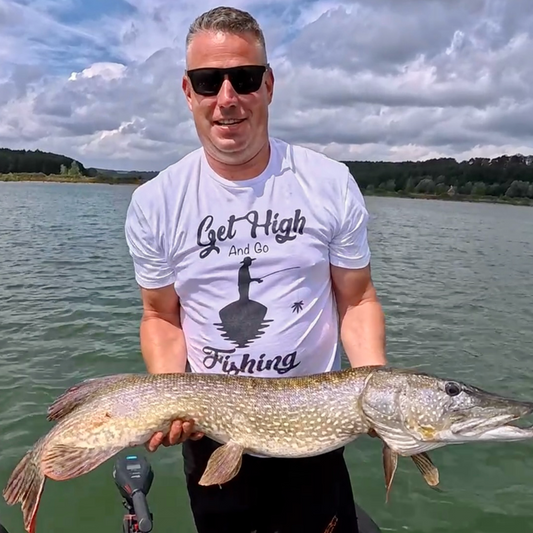 Angler auf dem Boot mit großem Hecht – trägt „Get HIGH and go FISHING“ T-Shirt, sonniger Tag auf dem See, Outdoor Fishing Motiv für Wels- und Raubfischangler